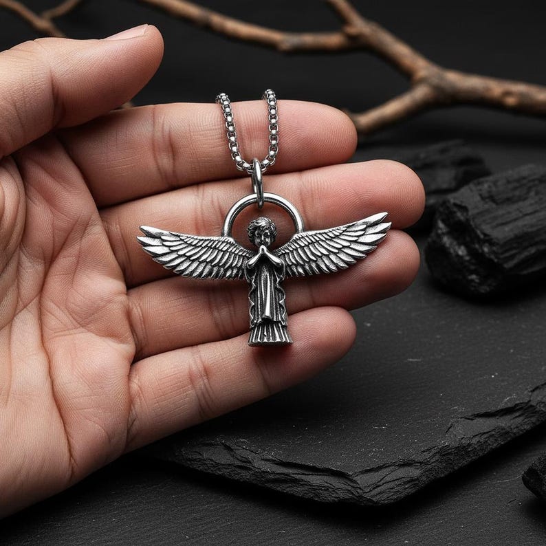 May include: Silver-toned angel pendant necklace. The pendant features an angel with outstretched wings and praying hands. The necklace has a chain with a circular bail. The angel pendant is held in a hand.