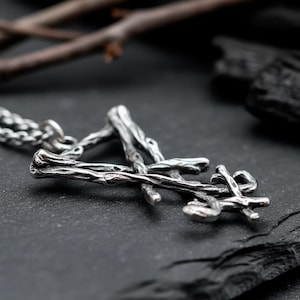 Puede incluir: Un collar con colgante plateado con un dise&ntilde;o similar a una ramita. El colgante est&aacute; hecho de ramas entrelazadas, creando una est&eacute;tica natural y org&aacute;nica. El collar se muestra sobre una superficie oscura y texturizada, con ramitas en el fondo.