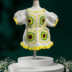May include: A white crochet dress featuring a square neckline and puff sleeves. The garment is constructed from multiple granny square motifs in white, yellow, lime green, and dark brown. The hem and sleeve edges are finished with yellow trim.