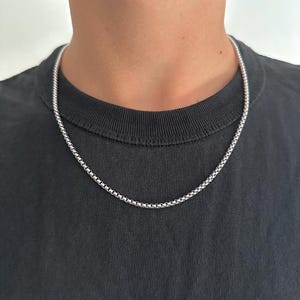 Op de afbeelding: Een zilverkleurige ketting met een vierkant schakelontwerp. De ketting wordt om de nek gedragen en contrasteert met een zwart T-shirt. De ketting is gemaakt van kleine, onderling verbonden schakels.