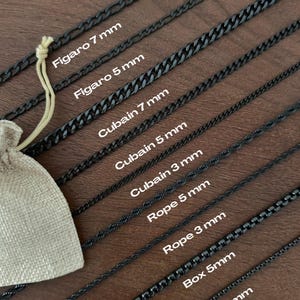 Pode incluir: Uma variedade de colares de corrente de metal preto em vários estilos, incluindo correntes Figaro, Cubana, Corda e Box. A imagem mostra diferentes larguras, com etiquetas indicando o tipo de corrente e o tamanho em milímetros. Uma pequena bolsa bege com cordão está na imagem.