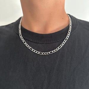 Pode incluir: Um colar de corrente Figaro prateado. O colar é feito de elos ovais e alongados interligados. A corrente é usada ao redor do pescoço, repousando sobre uma camiseta preta.