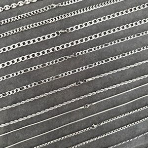 Pode incluir: Uma coleção de colares de corrente prateados em vários estilos, incluindo correntes de elos, figaro, corda e caixa. Cada corrente é exibida em uma superfície cinza, mostrando os diferentes designs de elos e detalhes de fecho.