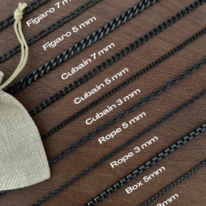 Pode incluir: Uma variedade de colares de corrente de metal preto em vários estilos e larguras, incluindo correntes Figaro, Cuban, Rope e Box. A imagem mostra os tamanhos das correntes em milímetros, variando de 3 mm a 7 mm, com um pequeno saco bege.