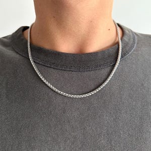 Può includere: Una collana a catena color argento con un design intrecciato. La collana è mostrata su una maglietta grigia. La catena è un accessorio semplice ed elegante.