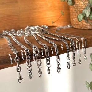 Può includere: Una collezione di collane a catena color argento esposte su una mensola di legno. Le catene variano nello stile, tra cui Coffee, Cuban, Gourmette, Figaro, Rope, Box, Spirale, Small Fine e Small Box. Ogni catena ha una chiusura.