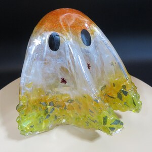 May include: A handcrafted ghost-shaped resin decoration. The top is orange, transitioning to clear with black eye cutouts. The bottom is yellow-green with embedded leaf-like shapes. Small red and silver leaf accents are scattered throughout.