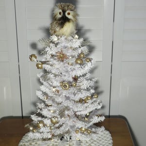 May include: A small, white Christmas tree decorated with gold ornaments and a decorative owl topper. The tree is set on a cream-colored tree skirt. The tree is on a wooden table.