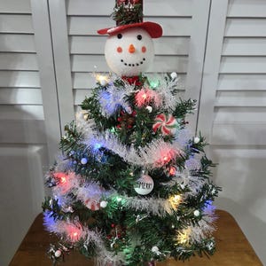 May include: A festive Christmas tree with a snowman topper. The snowman has a red hat and a smiling face. The tree is decorated with colorful lights, white garland, and a "Merry" ornament. Candy canes add to the holiday cheer.