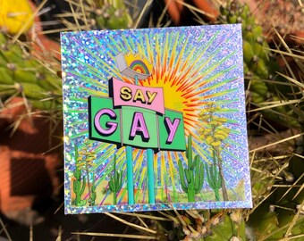 Puede incluir: Una pegatina holográfica con un letrero de estilo retro que dice "Say Gay" en letras rosas y verdes. El letrero está situado sobre un fondo de paisaje desértico con un sol amarillo brillante y un arcoíris.
