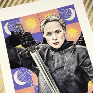 May include: A woman in silver armor holds a sword, standing in front of a colorful background with a repeating pattern of wolves and suns.