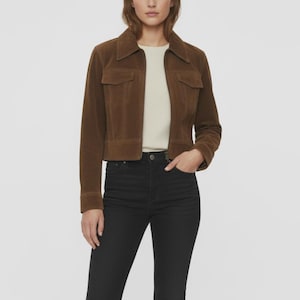 May include: A brown suede jacket with a collared neckline and two front pockets. The jacket is worn over a cream-colored top and paired with black jeans. The jacket has a zipper closure and long sleeves.