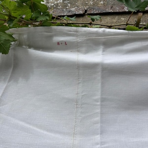 Puede incluir: Una tela textil blanca con una costura vertical visible que recorre el centro. El tejido muestra una textura sutil y presenta un pequeño monograma o marca bordada en rojo que consiste en dos caracteres cerca del borde superior.