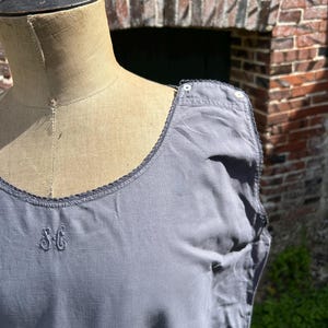 May include: A grey sleeveless garment with a scoop neckline, featuring a delicate dark grey lace trim along the edges. The fabric displays an embroidered monogram with the letters SG on the chest and two small white buttons on the shoulder.