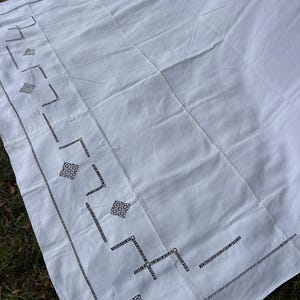 Può includere: Un tessuto rettangolare bianco caratterizzato da un bordo decorativo con motivi geometrici ricamati in nero e dettagli traforati. Il tessuto ha una consistenza liscia con cuciture visibili lungo i bordi e un motivo a rombi ripetuto.