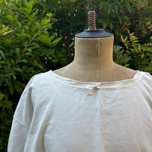 May include: A white linen shirt displayed on a beige mannequin. The shirt features a wide, rounded neckline with a small fabric tie closure at the center front. The fabric has a visible woven texture and a loose, relaxed fit with long sleeves.