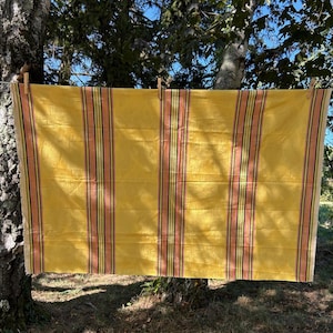 May include: A rectangular fabric piece in a vibrant yellow hue, featuring vertical stripes in shades of peach, green, and dark brown. The textile is suspended by three wooden clothes pegs, displaying a woven texture and clean, straight edges.