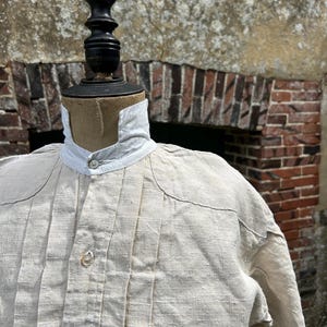 May include: A beige long-sleeved shirt featuring a white high collar with a single button closure, vertical pleats running down the front chest, and additional buttons along the centre placket, displayed on a black mannequin torso.