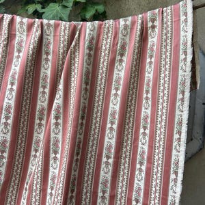 Peut inclure: Un coupon de tissu présentant un motif à rayures verticales rose poudré et blanc cassé. Le design alterne des bandes de rose uni et des bandes crème ornées de motifs répétitifs d'urnes fleuries dans des tons de rose et de vert.