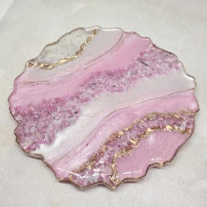 May include: A decorative coaster with a unique design. The coaster features a blend of pink and white hues, accented with gold glitter and a textured, irregular edge. The surface is embedded with small, sparkling fragments.