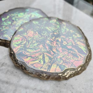 May include: Two iridescent coasters with a gold-colored trim. The coasters have a shimmering, multi-colored surface with hues of pink, green, and yellow. The edges have a rough, natural look.