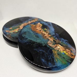 May include: Two round coasters with a glossy black finish. The coasters feature a swirling design of blue, green, and gold, with gold leaf accents. The coasters are designed to protect surfaces from drink rings.