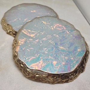 May include: Two iridescent, round coasters with a textured, gold-colored rim. The coasters have a shimmering, opalescent surface with hints of blue, pink, and yellow. These decorative coasters are designed to protect surfaces from drink rings.