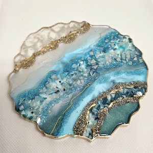 May include: A decorative coaster with a unique design. The coaster features a layered effect with shades of blue, teal, and white, accented with gold glitter and a gold trim. The surface is embedded with small, sparkling crystals.