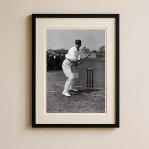 Puede incluir: Fotografía en blanco y negro enmarcada de un jugador de críquet en posición de bateo. El jugador viste uniforme blanco, gorra y equipo de protección. Un bate de críquet se sostiene listo para golpear. El fondo muestra un campo, espectadores y un edificio.