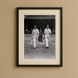May include: Framed black and white photograph of two cricket players walking on a field. They are wearing white uniforms, caps, and protective gear, carrying bats. The background shows a crowd and a white picket fence.