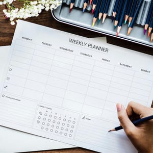 Peut inclure: Un planificateur hebdomadaire blanc avec le texte "WEEKLY PLANNER" est posé sur une surface en bois. Une main tient un crayon, prête à écrire. Des crayons de couleur sont dans un récipient en métal en arrière-plan. Des fleurs blanches sont également en arrière-plan.