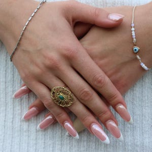 Könnte beinhalten: Nahaufnahme von Händen mit Schmuck. Ein goldener Ring mit einem grünen Stein sitzt an einem Finger. Ein silbernes Armband und ein zartes Armband mit einem bösen Blick-Anhänger sind ebenfalls zu sehen. Die Nägel sind mit einer weißen Spitze lackiert.