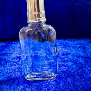 Pode incluir: Uma garrafa de vidro transparente e retangular com tampa prateada. A garrafa está vazia e sobre uma superfície de veludo azul. A tampa é cilíndrica e brilhante.