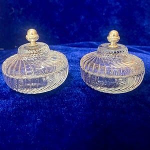 May include: Two clear glass trinket jars with spiral detailing and gold-colored finials. The jars are round with a lid and sit on a dark blue surface. The jars are likely vintage or antique.