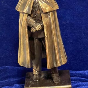 May include: Bronze statue of a man in military uniform, wearing a cape and hat. The statue is mounted on a rectangular base. The figure's hands are clasped in front of him. The statue has a detailed, aged appearance.
