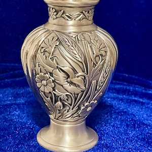 Pode incluir: Um vaso prateado com intrincadas esculturas florais em relevo. O vaso apresenta uma borda alargada e um corpo arredondado, adornado com representações detalhadas de flores, folhas e espigas de trigo. A base é um pedestal circular simples. O fundo é azul escuro.