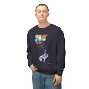 May include: A dark navy blue crewneck sweatshirt featuring a graphic print of an astronaut floating while holding onto a cluster of colorful planets tied together with strings, with the text "let go" printed in small white letters below the astronaut.