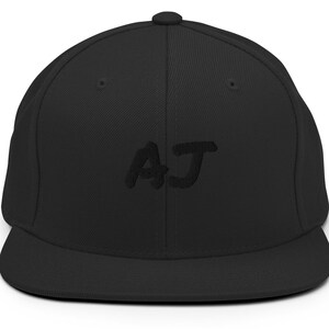 May include: A black, flat-brimmed baseball cap with the embroidered letters "AJ" in a matching black thread. The cap has a structured crown and a curved brim, suitable for casual wear.