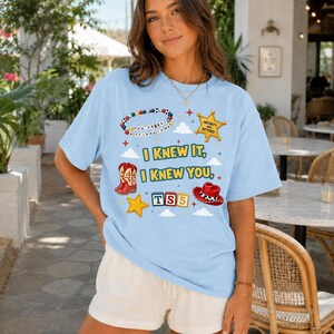 May include: A light blue t-shirt featuring a graphic print with the text "I KNEW IT, I KNEW YOU," "TS5," "INFINITY AND BEYOND," and "You've got a friend in me." The design includes illustrations of a beaded bracelet, a sheriff badge, cowboy boots, a hat, and blocks.