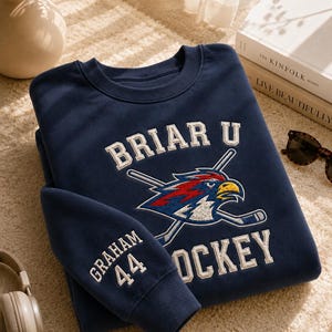 Op de afbeelding: Marineblauw sweatshirt met de tekst "BRIAR U HOCKEY" en een vogel logo met gekruiste hockeysticks. De mouw heeft de tekst "GRAHAM 44". Het sweatshirt ligt op een beige tapijt.