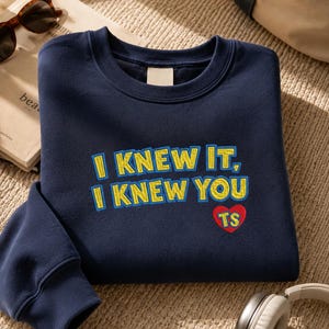 May include: A navy blue crewneck sweatshirt featuring embroidered text that reads I KNEW IT, I KNEW YOU in yellow with blue outlines. Below the text is a red heart shape with the letters TS embroidered in yellow inside the heart.