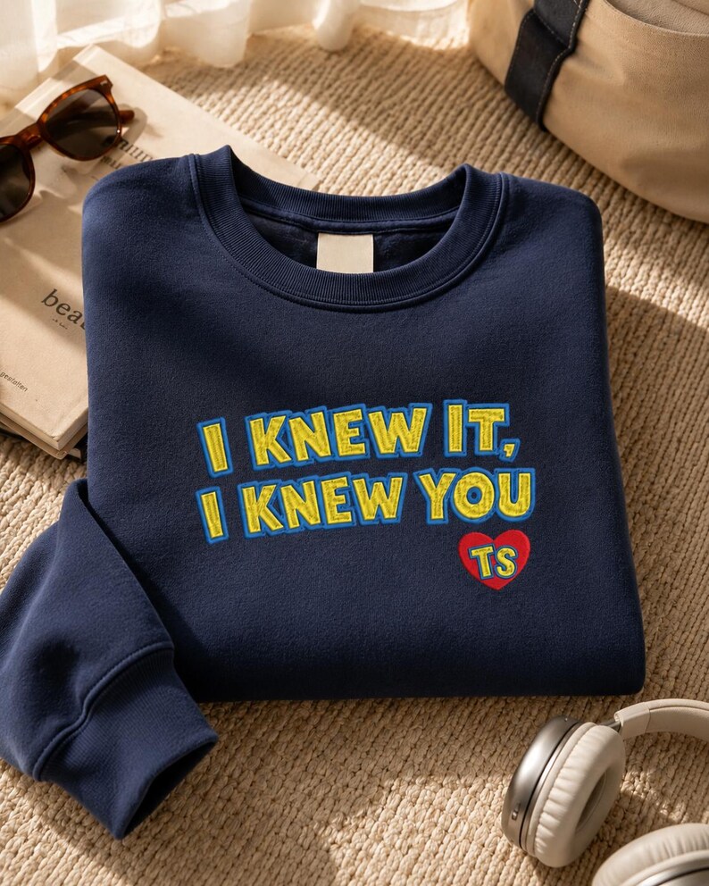 May include: A navy blue crewneck sweatshirt featuring embroidered text that reads I KNEW IT, I KNEW YOU in yellow with blue outlines. Below the text is a red heart shape with the letters TS embroidered in yellow inside the heart.