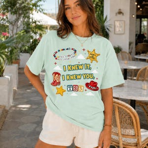 May include: A light green short-sleeve t-shirt featuring a graphic print with the text "I KNEW IT, I KNEW YOU," a beaded bracelet, a sheriff badge, red cowboy boots, a yellow star, "TS5" blocks, and a red cowboy hat with white trim.