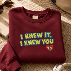 May include: A maroon crewneck sweatshirt featuring embroidered text in yellow with a blue outline that reads I KNEW IT, I KNEW YOU. Below the text is a small red heart shape containing the yellow embroidered letters TS on the front chest area.