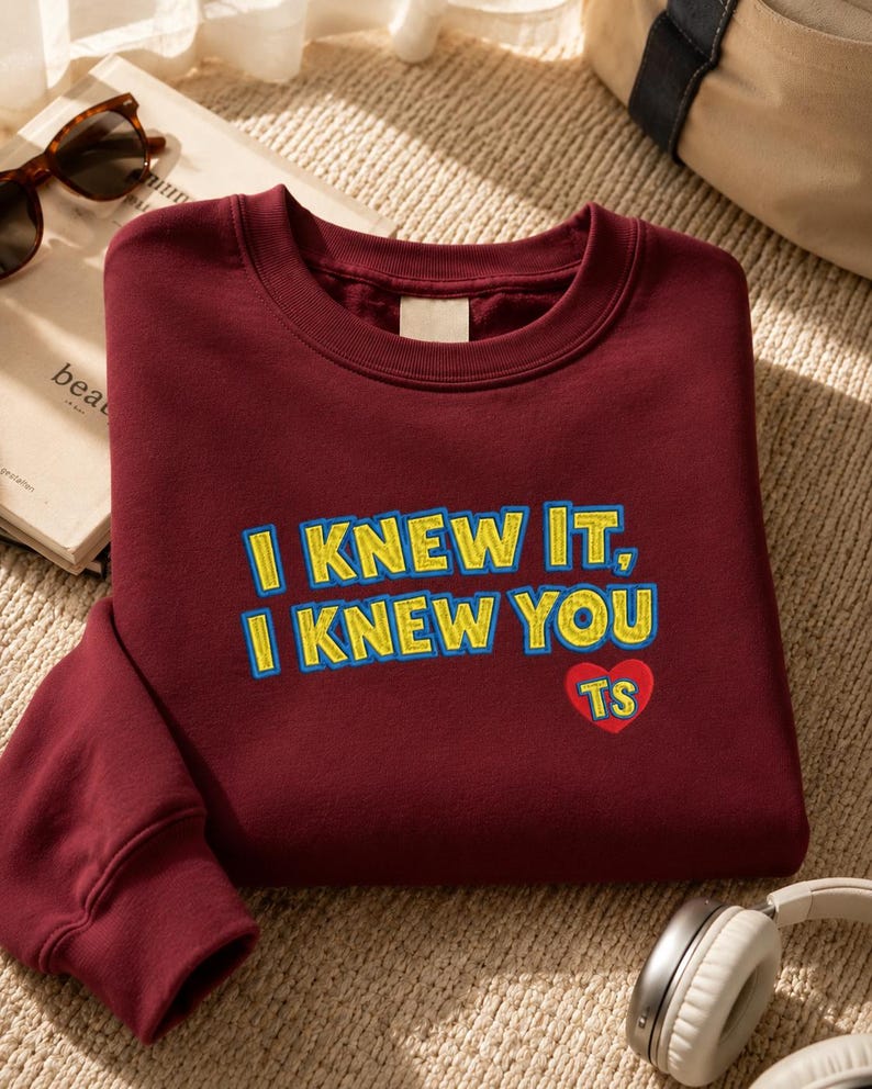 May include: A maroon crewneck sweatshirt featuring embroidered text in yellow with a blue outline that reads I KNEW IT, I KNEW YOU. Below the text is a small red heart shape containing the yellow embroidered letters TS on the front chest area.