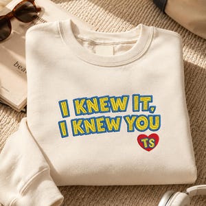 May include: A cream-colored crewneck sweatshirt featuring embroidered text that reads I KNEW IT, I KNEW YOU in yellow with blue outlines. Below the text is a red heart shape with the letters TS embroidered in yellow inside the heart.