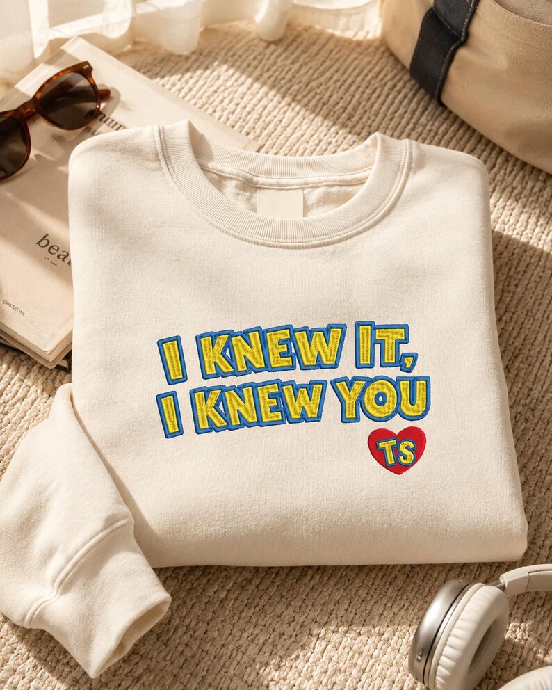 May include: A cream-colored crewneck sweatshirt featuring embroidered text that reads I KNEW IT, I KNEW YOU in yellow with blue outlines. Below the text is a red heart shape with the letters TS embroidered in yellow inside the heart.