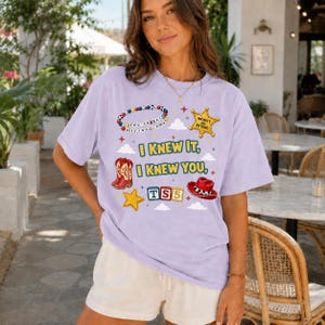 May include: A light purple short-sleeve t-shirt featuring a graphic print with a beaded necklace, a sheriff star badge, red cowboy boots, alphabet blocks spelling TS5, and a red cowboy hat, with the text I KNEW IT, I KNEW YOU.