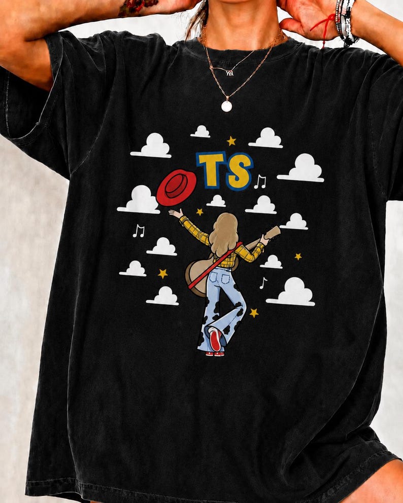 Peut inclure: Un t-shirt noir avec un motif repr&eacute;sentant une personne aux cheveux longs portant une chemise &agrave; carreaux jaune et un jean pattes d'eph &agrave; motif vache, tenant une guitare et un chapeau rouge. Le design inclut des nuages blancs, des &eacute;toiles jaunes, des notes de musique et les lettres TS.