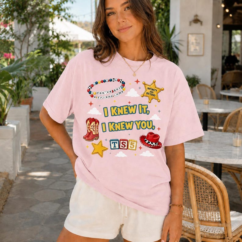 May include: A light pink short-sleeve t-shirt featuring a graphic print with a beaded bracelet, a sheriff badge, cowboy boots, a cowboy hat, and alphabet blocks. The text reads: You've got a friend in me, Infinity and beyond, I KNEW IT, I KNEW YOU, TS5.